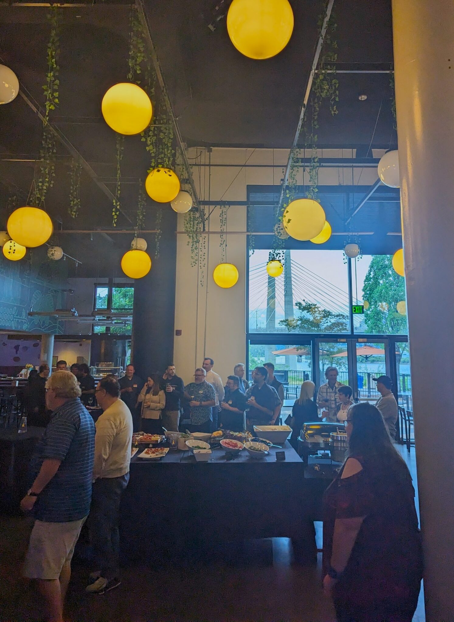 Shot of SpireTech's 30th anniversary at OMSI: People gathered, hanging lights, and a view of a Portland bridge beyond. 