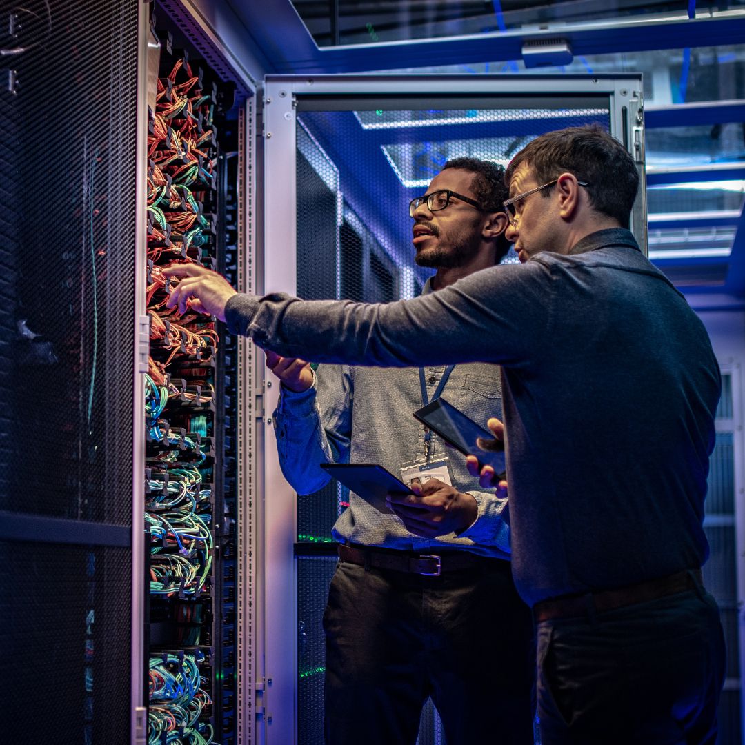 2 professionals standing in front of a computer server with lots of wires.